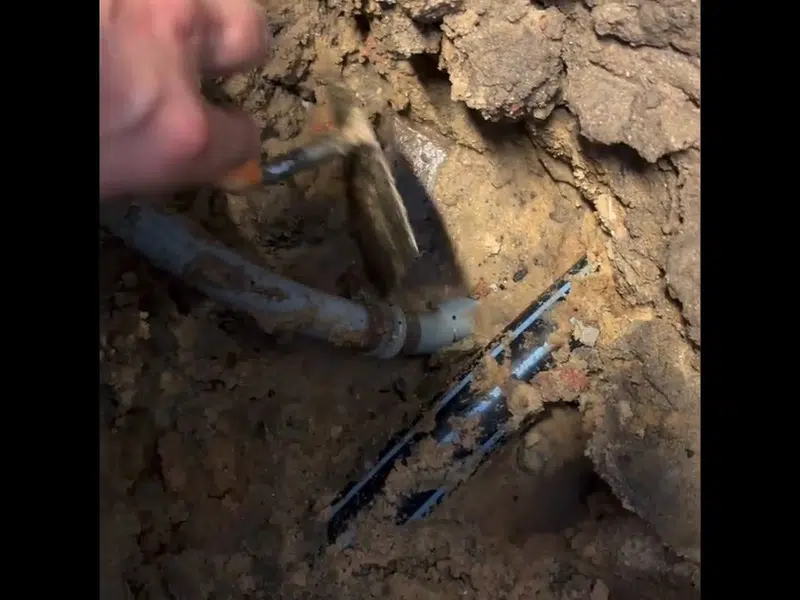 Hand exposing broken pipe in dirt trench for burst pipe repair in Stanton